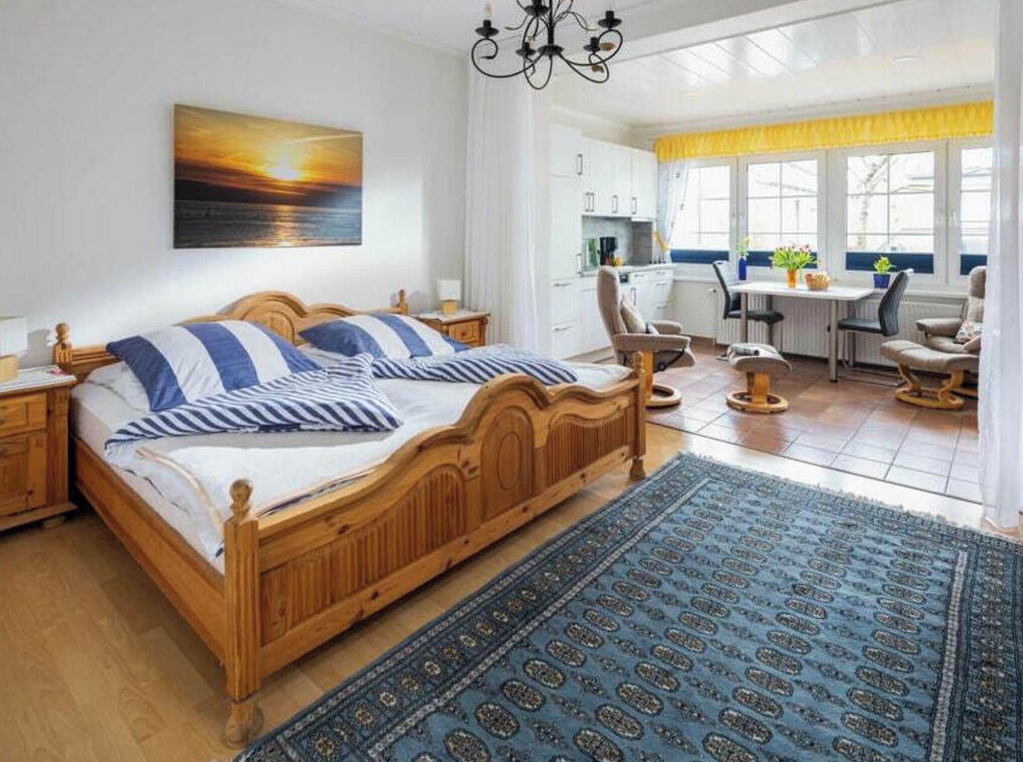 A bedroom with a wooden bed, a blue patterned rug, a painting of a sunset, and a view of the kitchen and dining area.