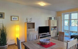 Ein Wohnzimmer mit Fernseher, einem Esstisch mit Stühlen, einem Bücherregal und einem Fenster mit Jalousien.