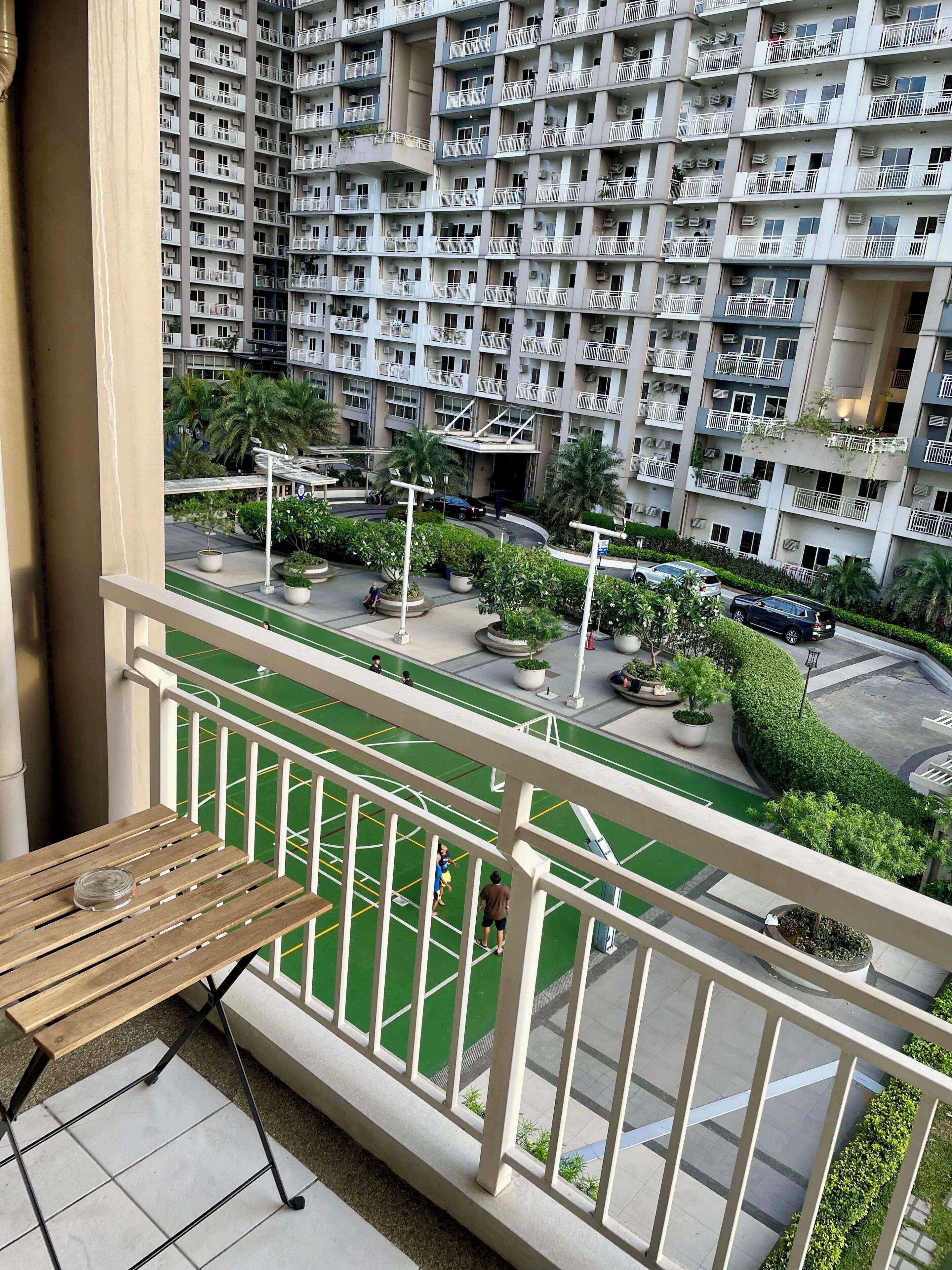 A balcony with a view of a residential building, a green sports court, and a parking area.