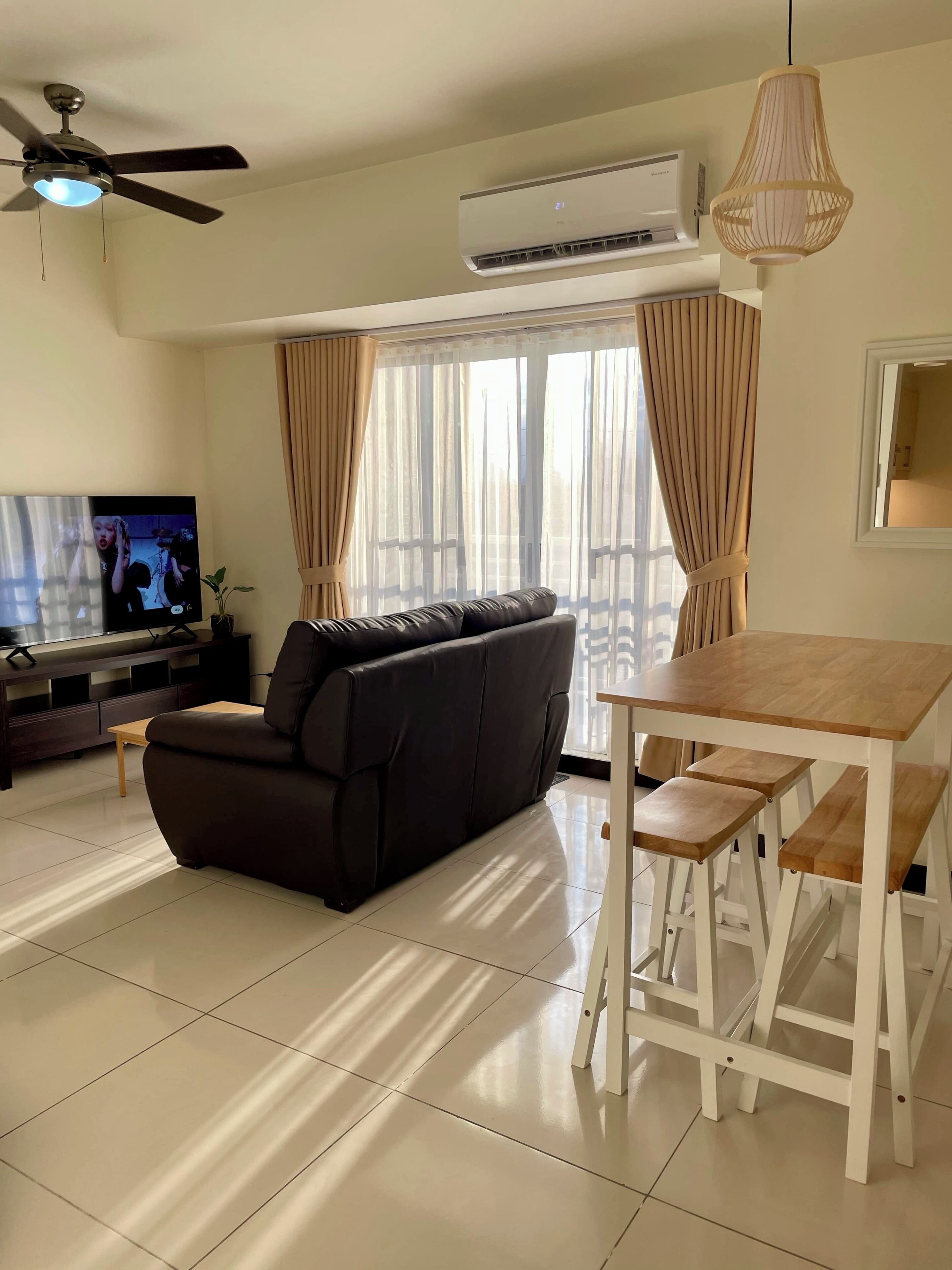 A modern living room with a black leather sofa, a wooden dining table with benches, a ceiling fan, and an air conditioning unit.