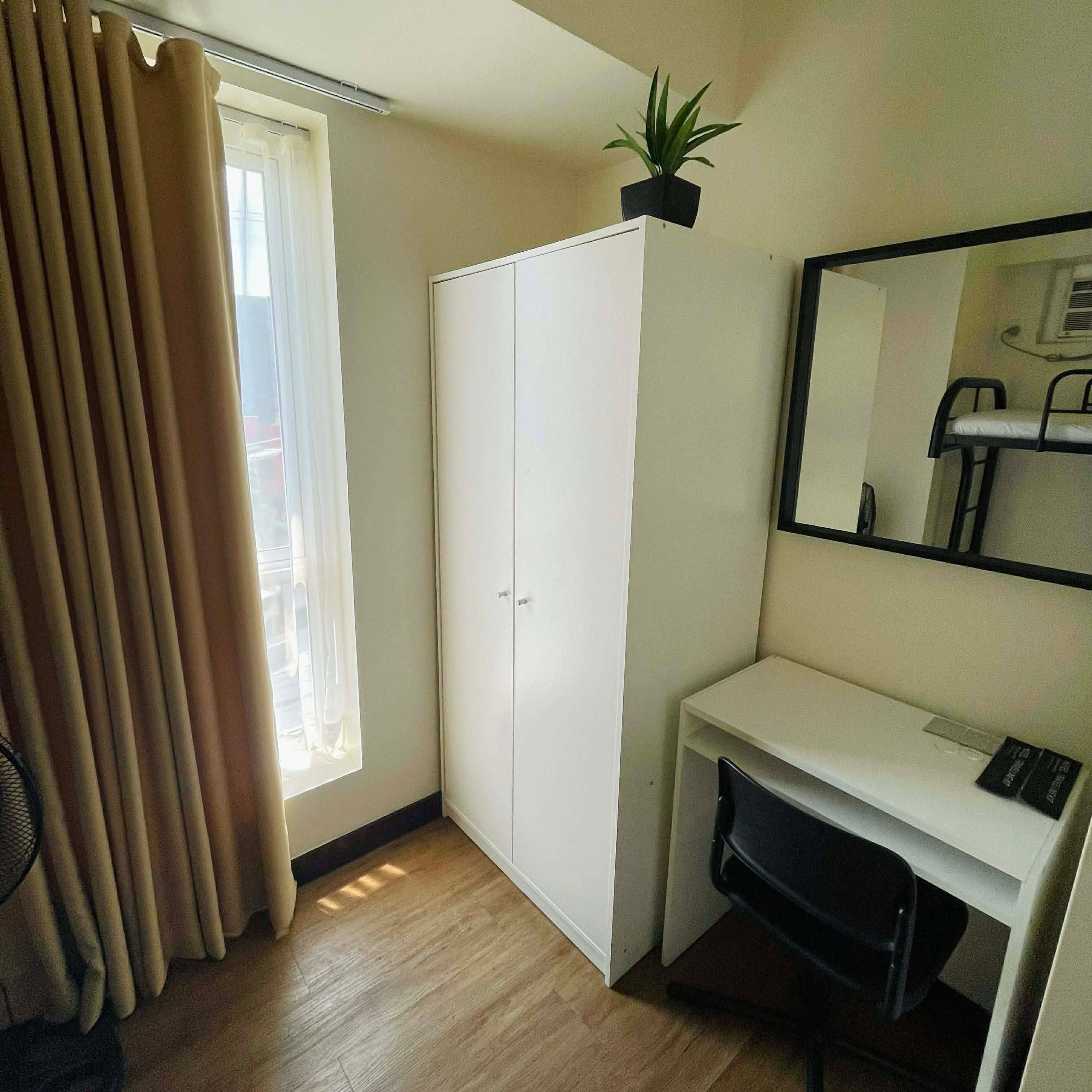 A room with a wardrobe, a desk, a chair, a potted plant, and a window with curtains.