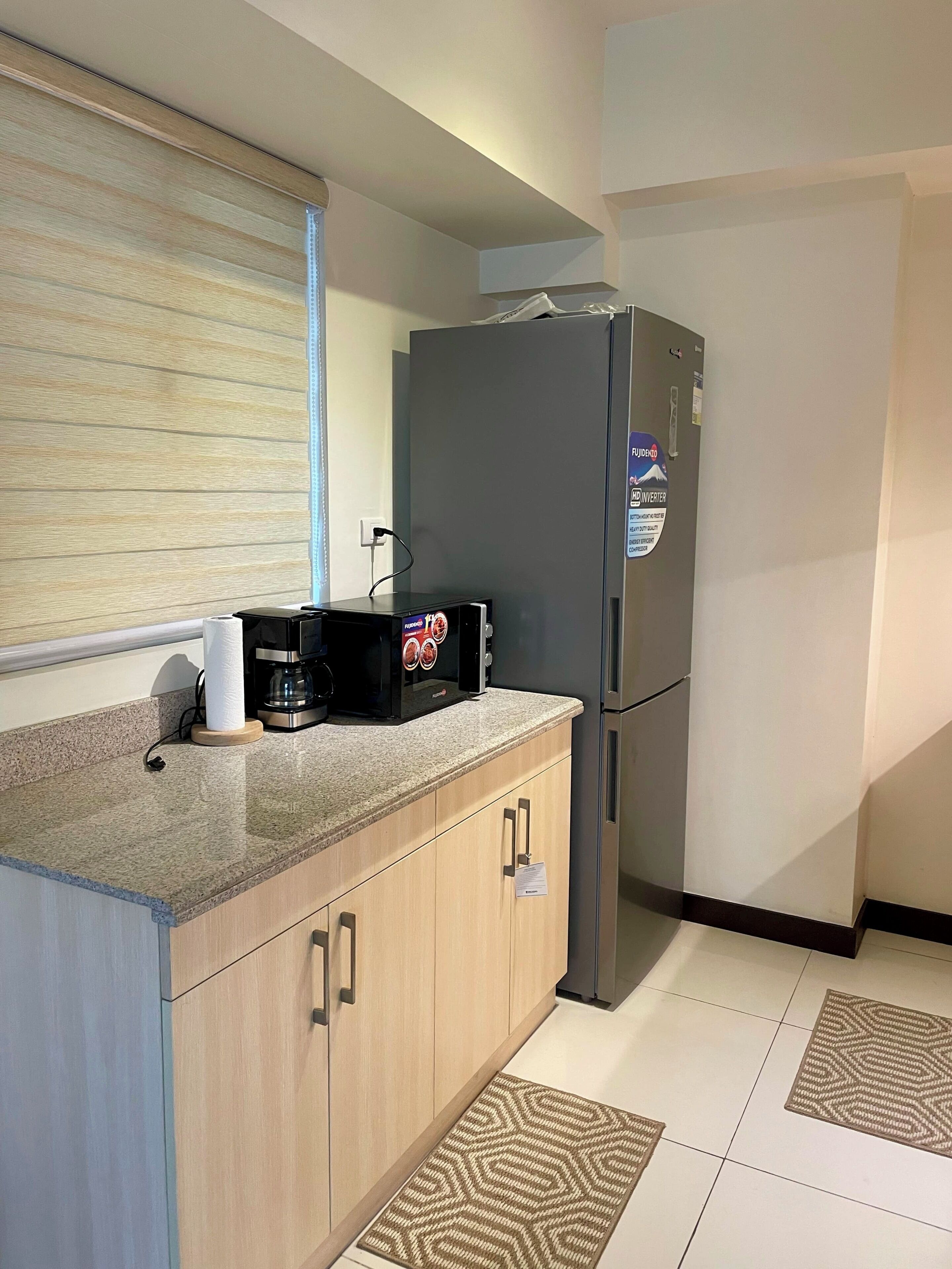 Une cuisine compacte équipée d’un réfrigérateur, d’une machine à café et d’une fenêtre avec des stores.