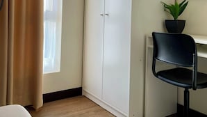 Ein Zimmer mit einem Bett, einem Kleiderschrank, einem Schreibtisch mit Stuhl, einer Zimmerpflanze und einem Fenster mit Vorhängen.