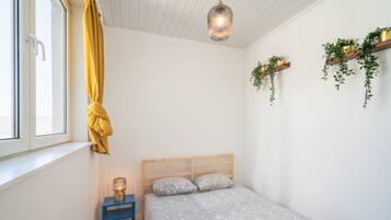 Quarto com cama, mesa de cabeceira, abajur, janela com cortinas e plantas na parede.