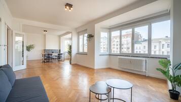 Amplia sala de estar con piso de madera, sofá, mesa de comedor con sillas y ventanas grandes que ofrecen vistas de edificios.