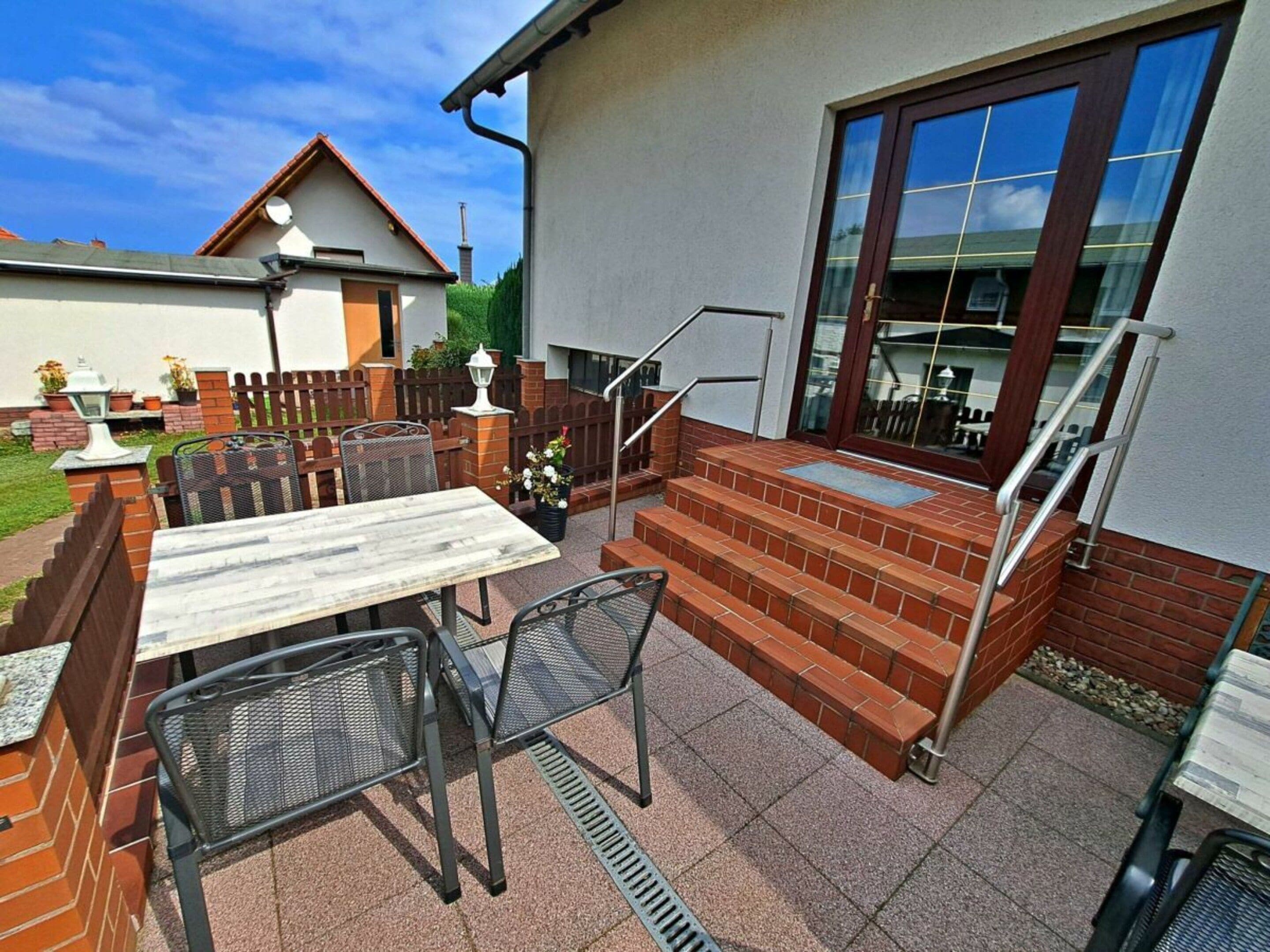 Een terras buiten met een tafel, stoelen, een meten trap en een glazen deur die leidt naar een overdekt gedeelte.