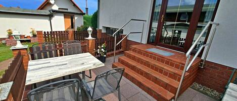 Een terras buiten met een tafel, stoelen, een meten trap en een glazen deur die leidt naar een overdekt gedeelte.
