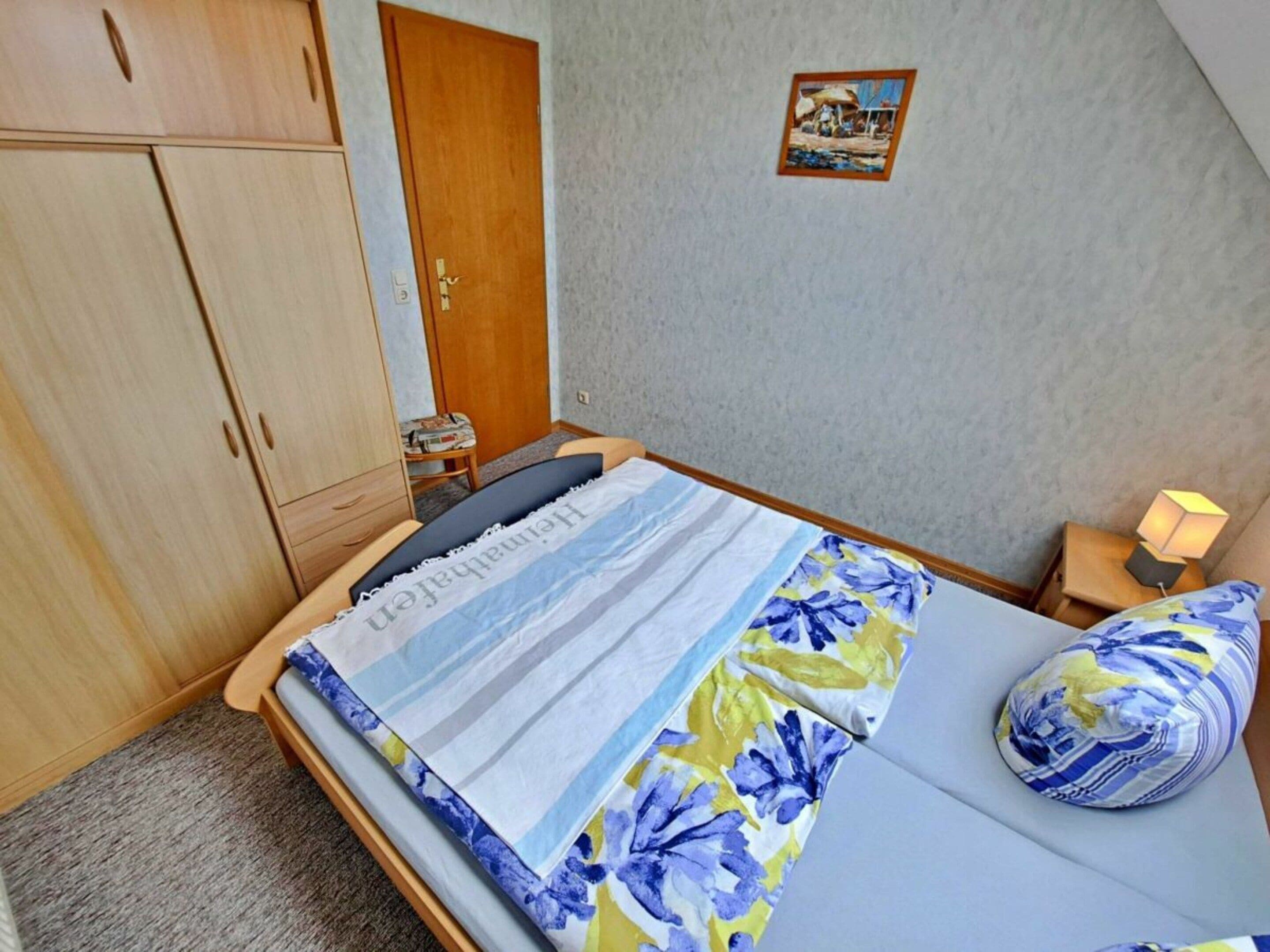 A bedroom with a bed, a nightstand, a lamp, a wardrobe, and a picture on the wall.
