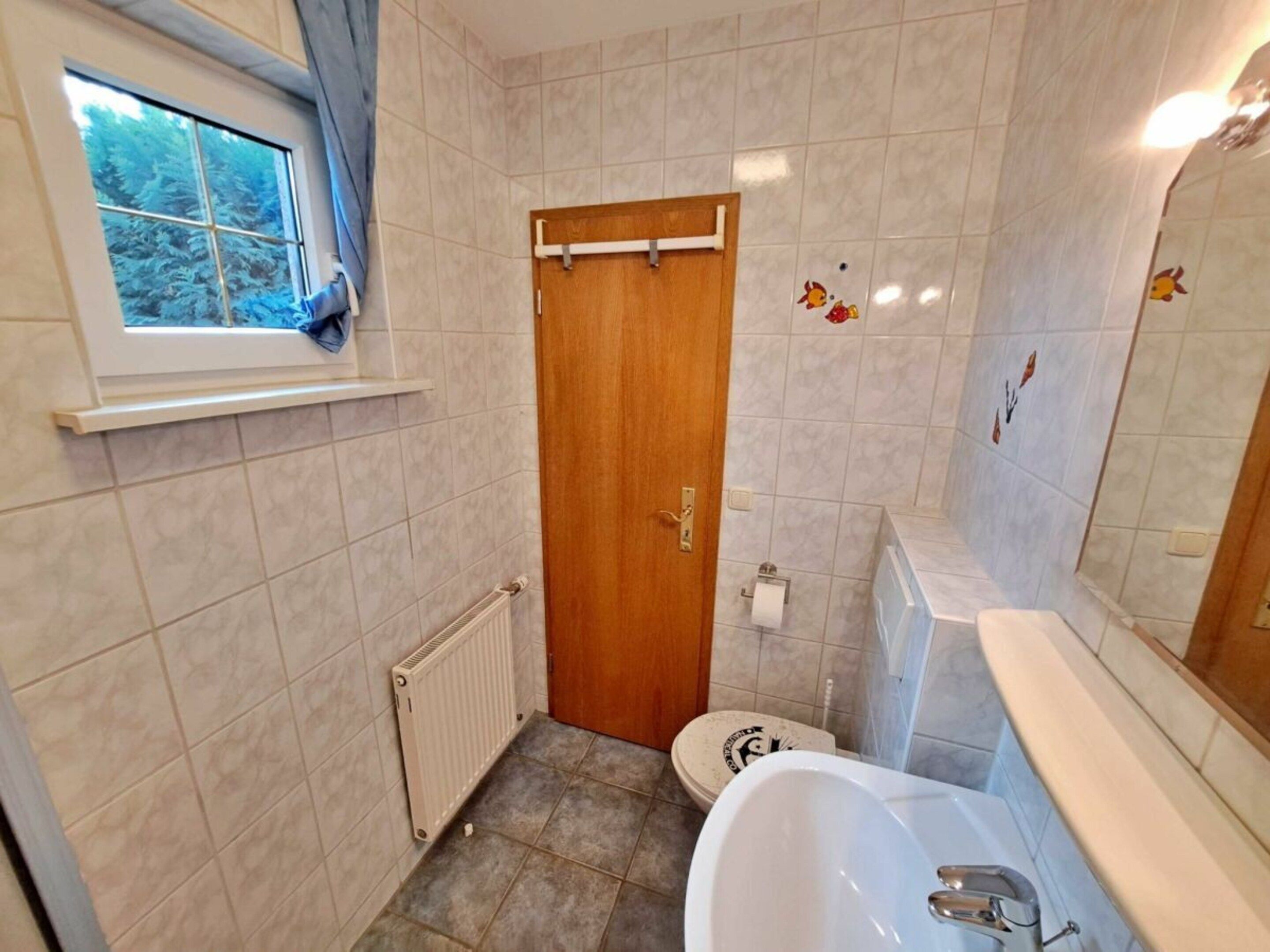 Un baño con puerta de madera, inodoro, bidé, radiador y ventana con vista a árboles.