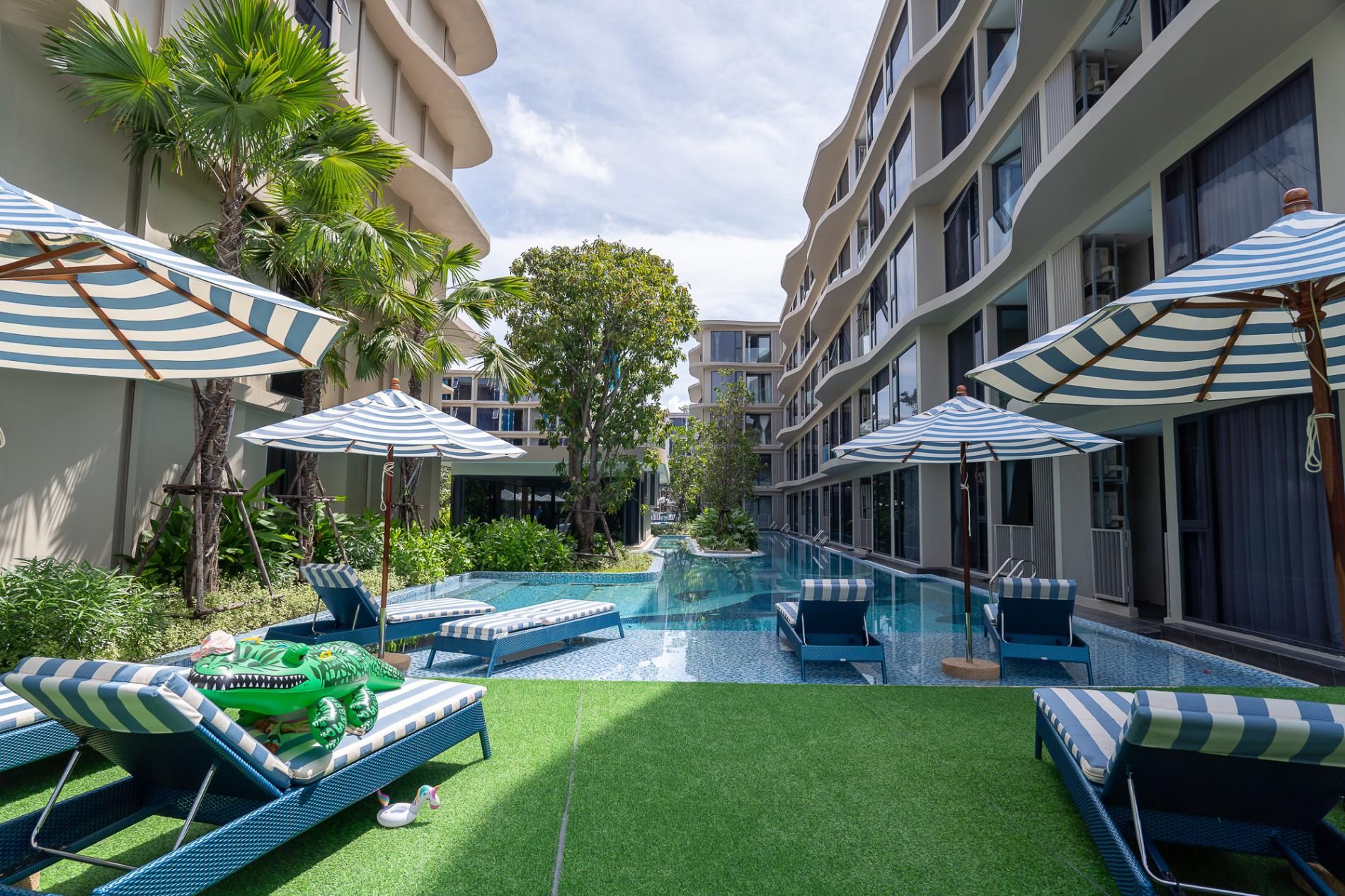 A hotel pool area with lounge chairs and umbrellas, surrounded by greenery and modern architecture.
