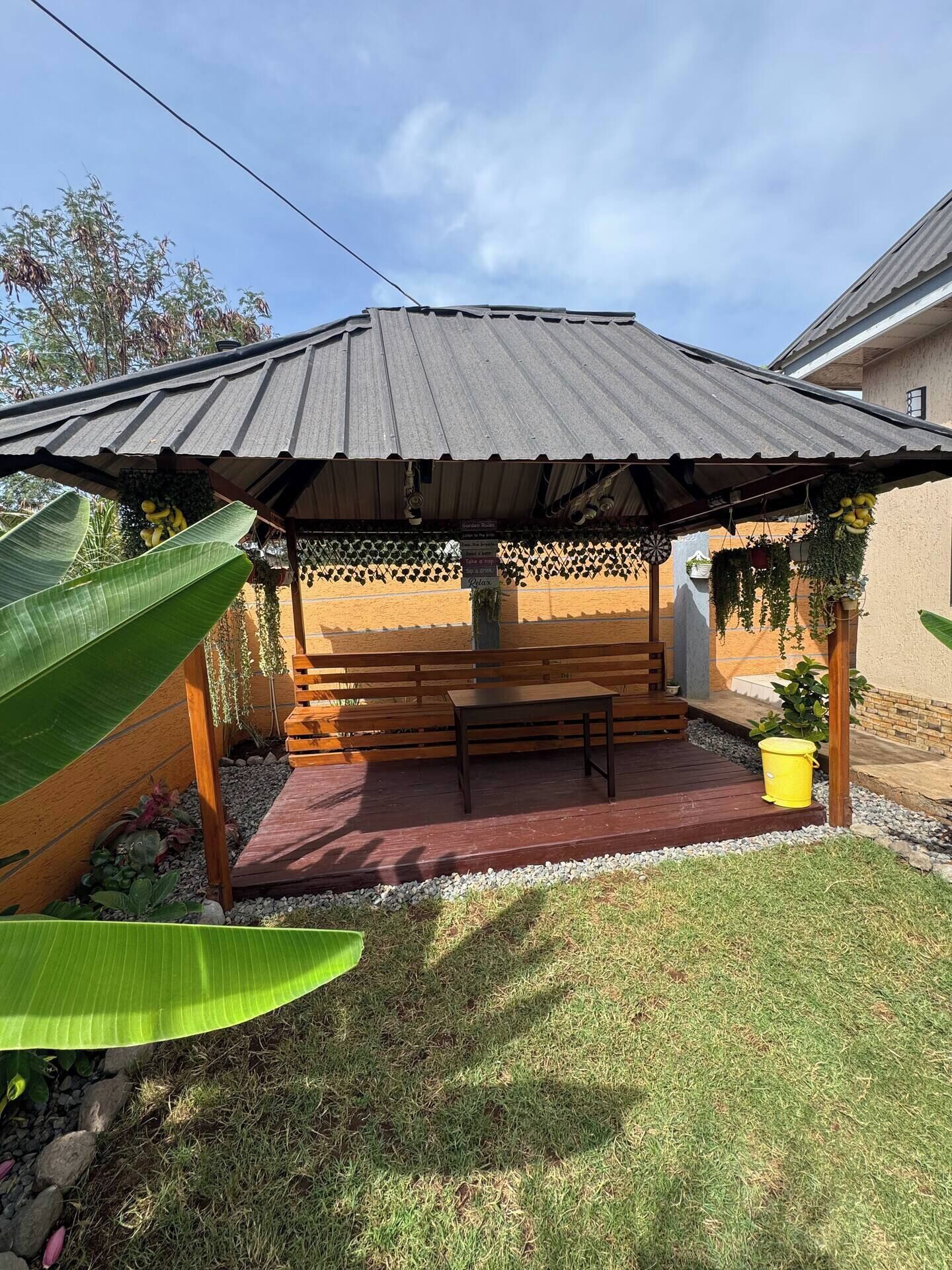 A covered outdoor seating area with a bench and a table, surrounded by plants and a grassy lawn.