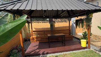 A covered outdoor seating area with a bench and a table, surrounded by plants and a grassy lawn.