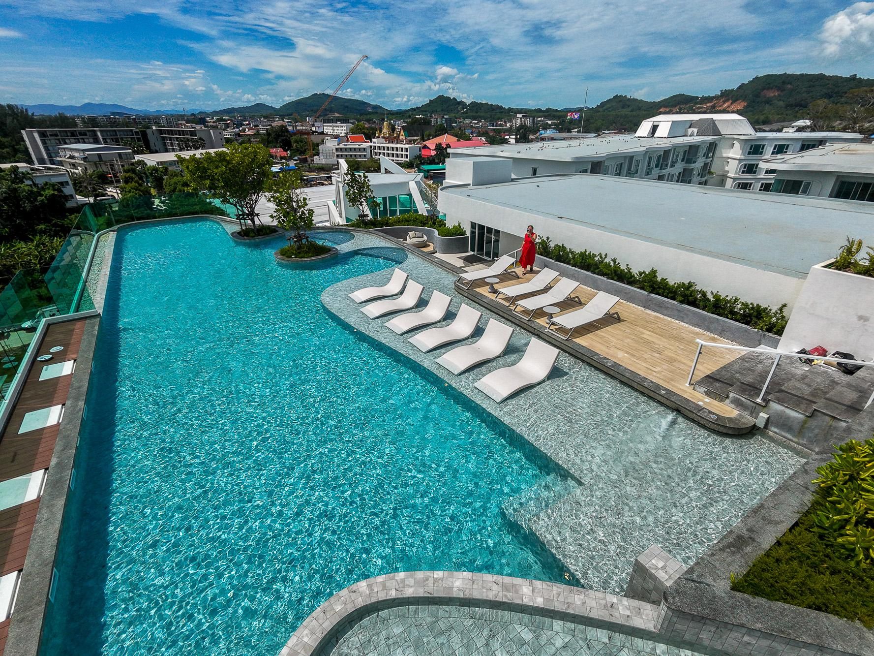 A large swimming pool with lounge chairs, a view of the cityscape, and a mountainous backdrop.