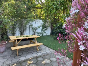 A wooden picnic table with benches on a paved patio surrounded by greenery and a white fence. - Serenity Now Guest House (Los Angeles)