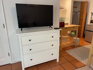 Een witte dressoir met een flatscreen televisie erop, een houten bijzettafeltje met een mand, en een betegelde vloer.