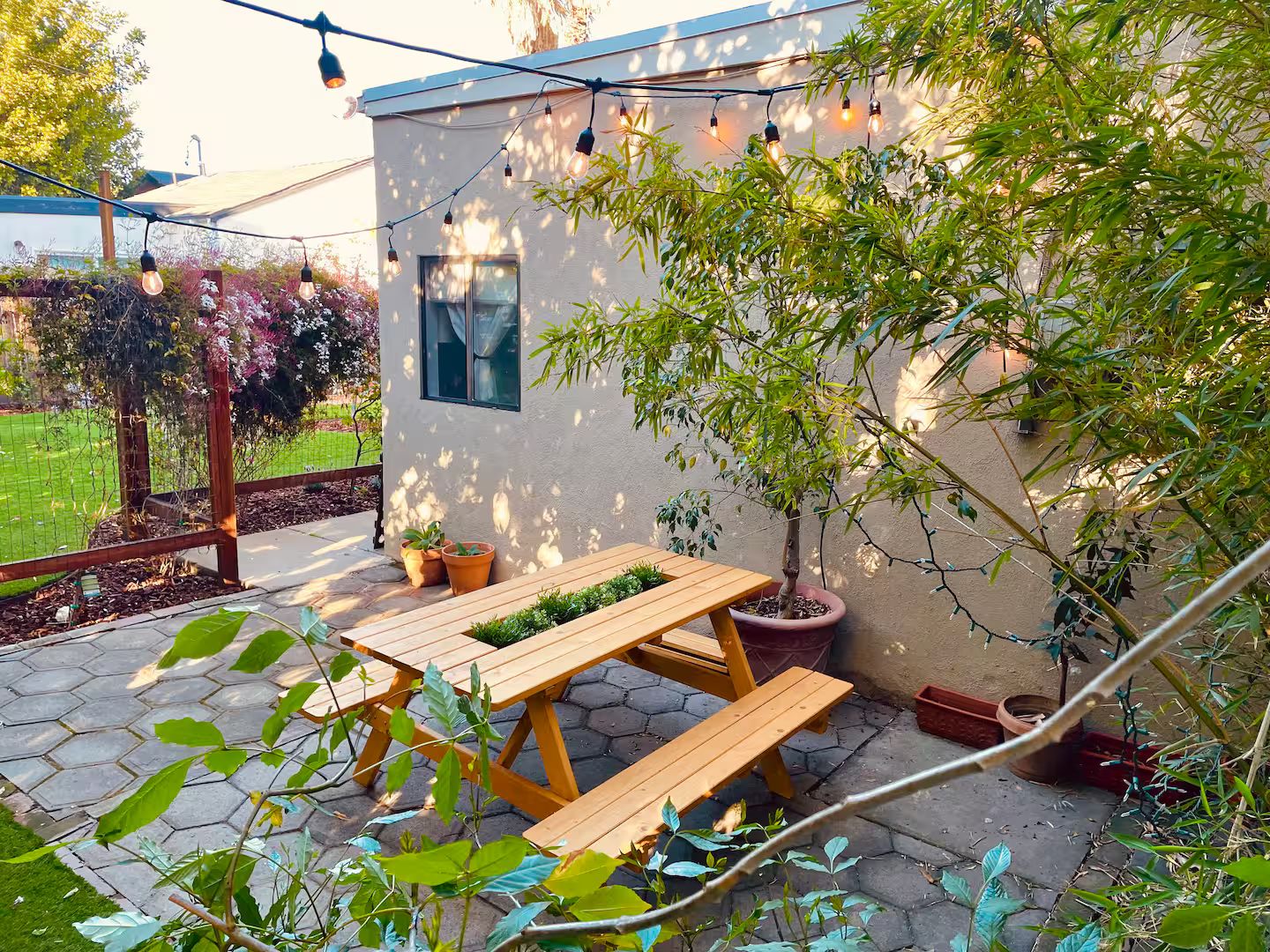 Een terras met houten banken en een tafel, omringd door potplanten en een lichtslingering.