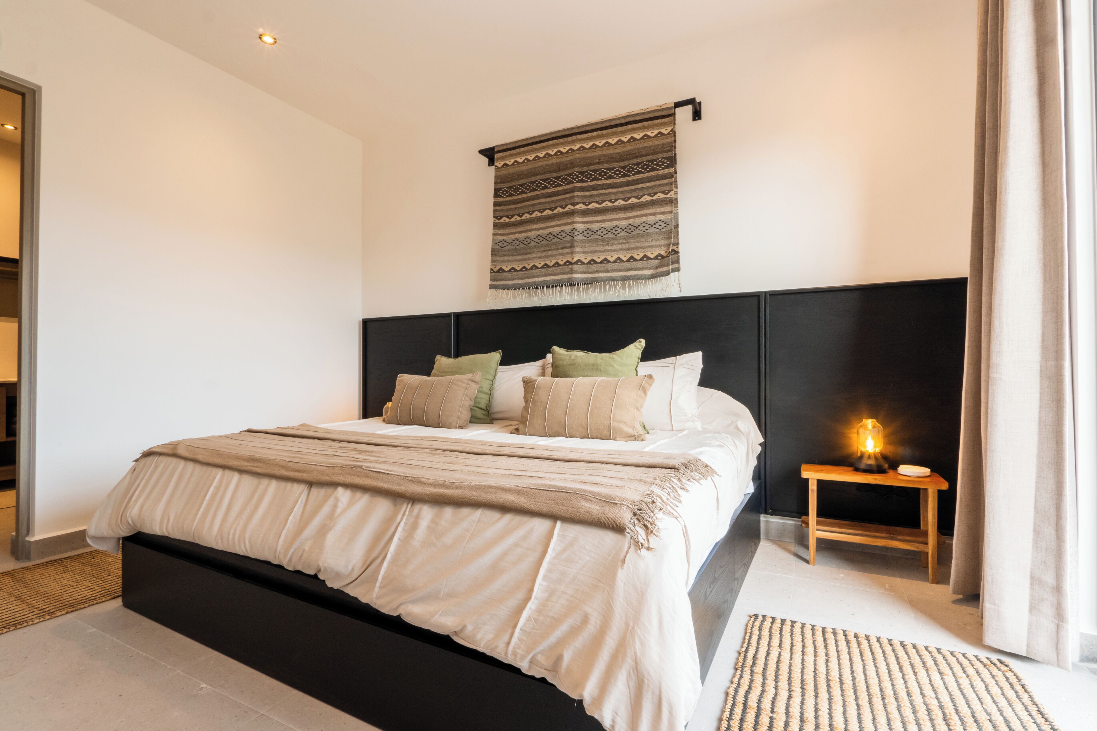 A bedroom with a large bed, a headboard, a bedside table with a lamp, and a patterned rug on the floor.