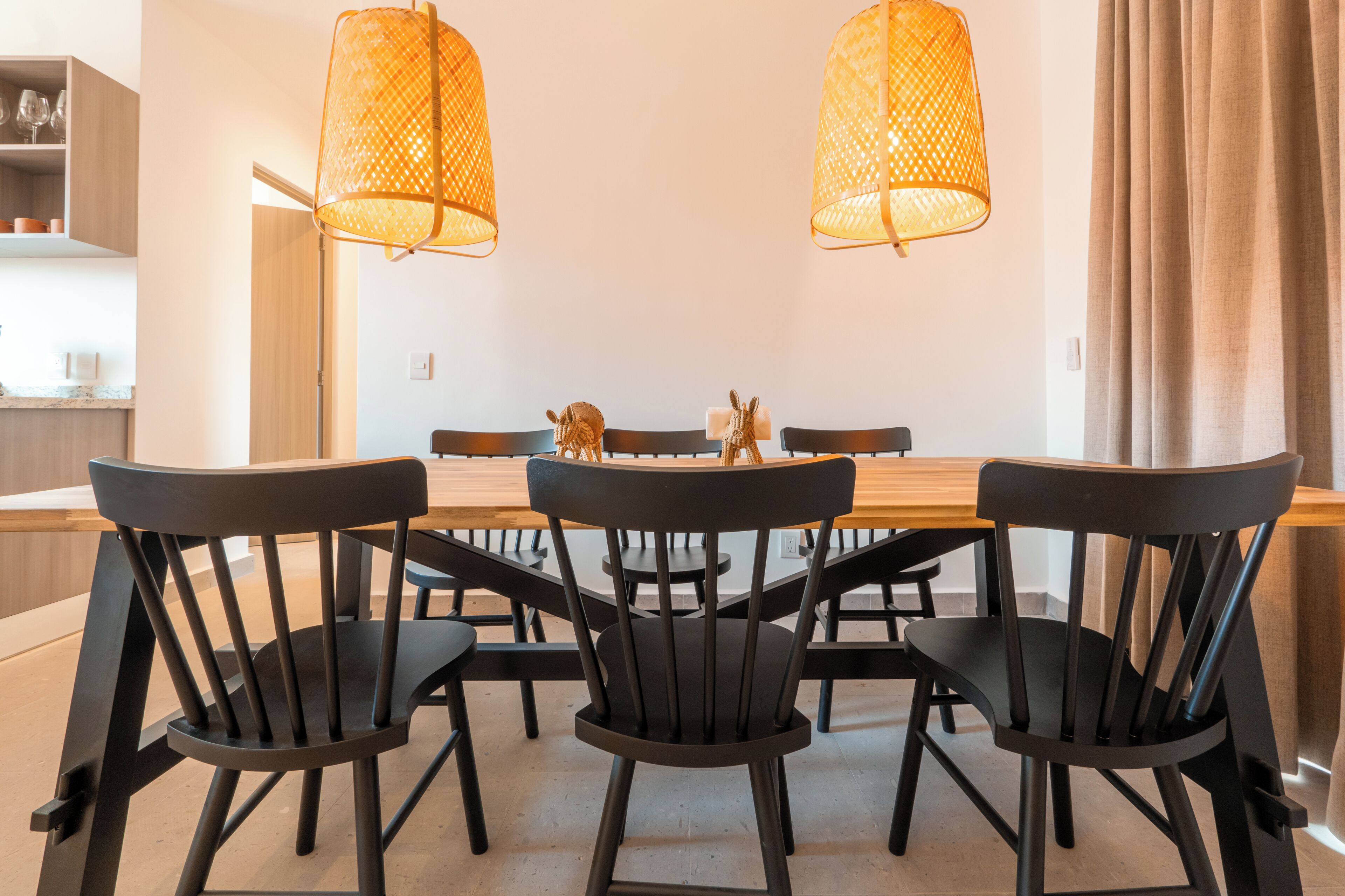 A dining area with a wooden table and black chairs, two hanging lamps, and a kitchen area in the background.