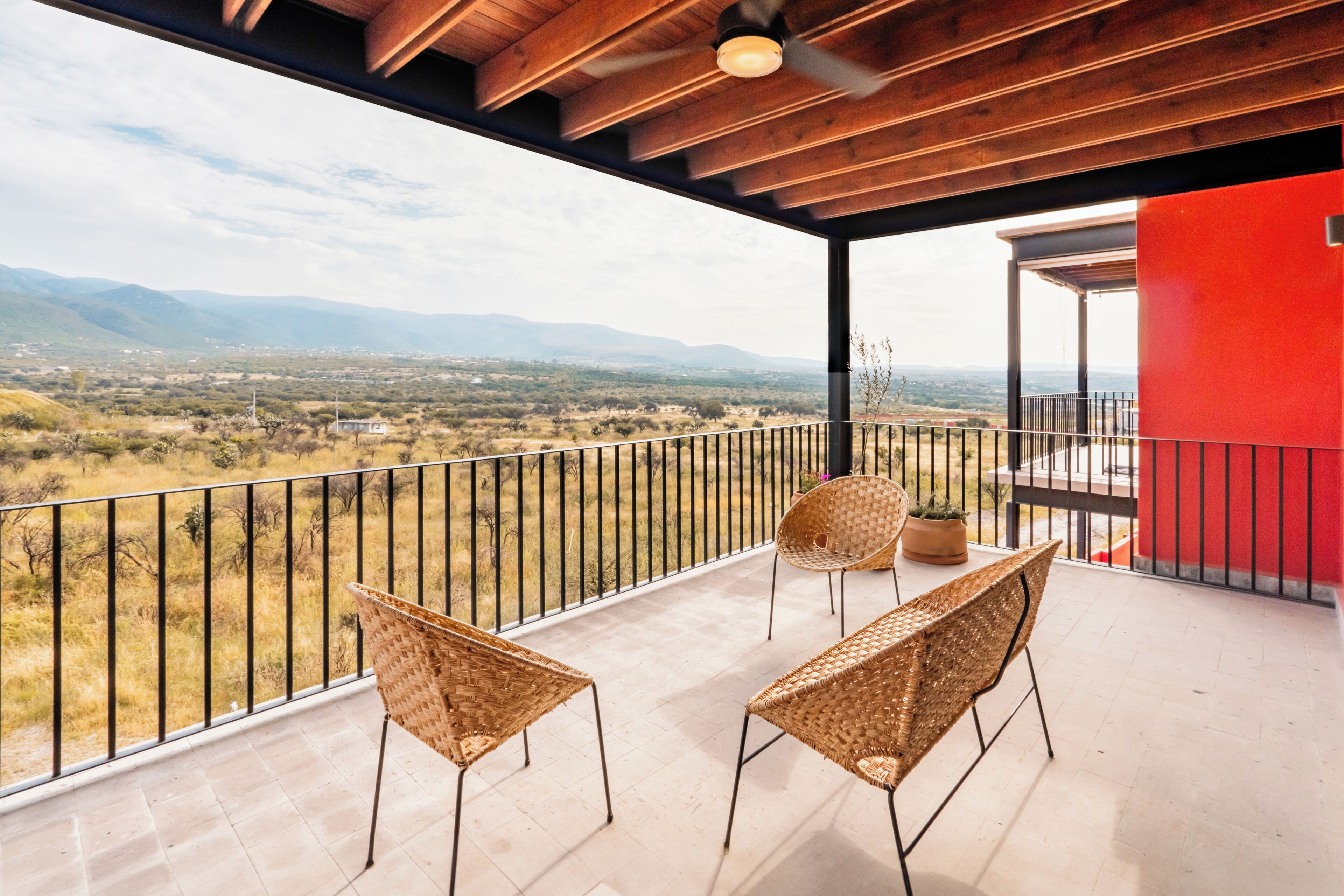 A balcony with two wicker chairs, a potted plant, and a view of a landscape with mountains in the distance.