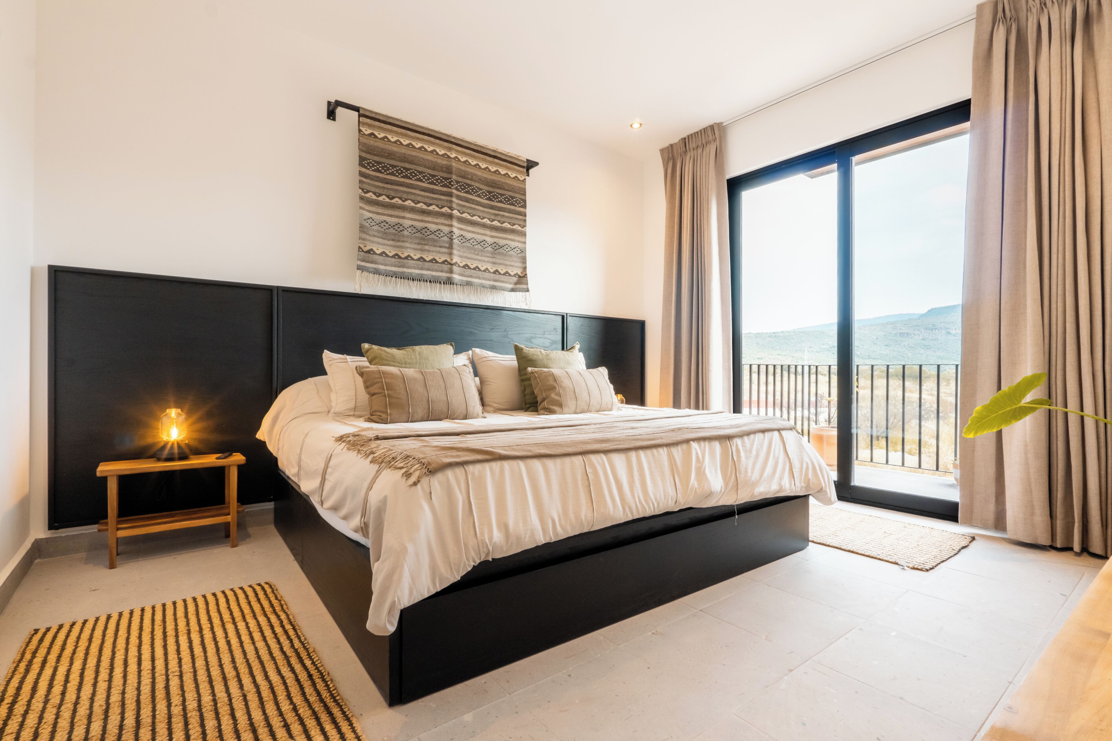 A modern bedroom with a large bed, a nightstand, a wall-mounted lamp, and a view of the outdoors through a sliding glass door.