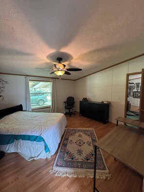 A bedroom with a bed, a desk, a chair, a ceiling fan, and a window with a view of a car outside. - Old but bright & comfy modular home with wildlife on serene acre; family & pets (Lanesville)