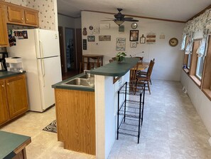 A kitchen with wooden cabinets, a refrigerator, a sink, and a dining area with a table and chairs. - Old but bright & comfy modular home with wildlife on serene acre; family & pets (Lanesville)