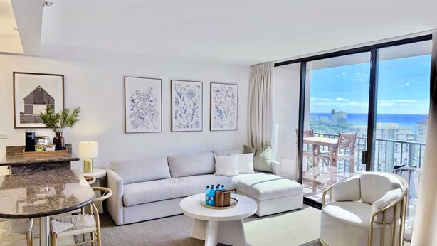 Ein modernes Wohnzimmer mit einer Couch, einem runden Couchtisch und einem Sessel. An der Wand hängen gerahmte Pflanzenbilder und durch große Fenster ist die Stadtansicht zu sehen.
