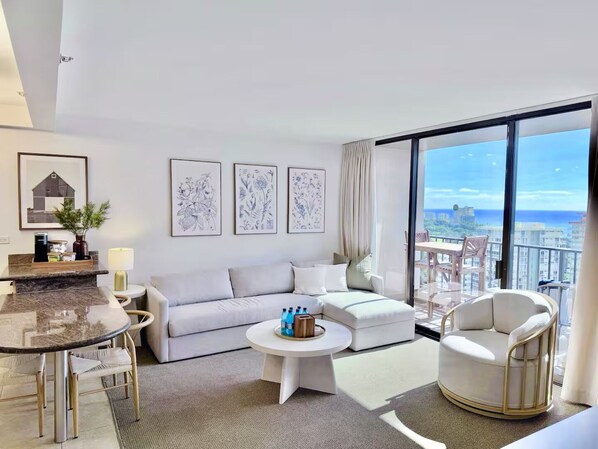 A modern living room with a sofa, a round coffee table, and a chair. There are framed botanical prints on the wall and a view of the city through large windows. - 23F-Waikiki Gem-1BR Condo with Diamond Head & Ocean Views (Honolulu)
