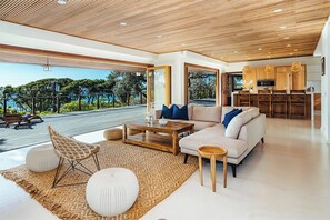 A modern living room with a sofa, coffee table, and chairs, a kitchen area, and a view of the outdoors. - Diamond Head Tranquility: 4BR Ocean View Home (Honolulu)