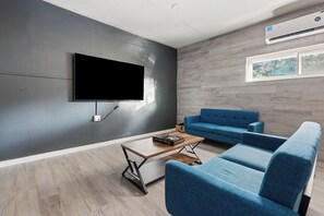 A modern living room with a flat-screen TV mounted on a grey wall, a blue sofa, a wooden coffee table, and a small bookshelf. - Yosemite Gateway Oasis - A Spacious 7 Bedroom house with pool (Oakhurst)