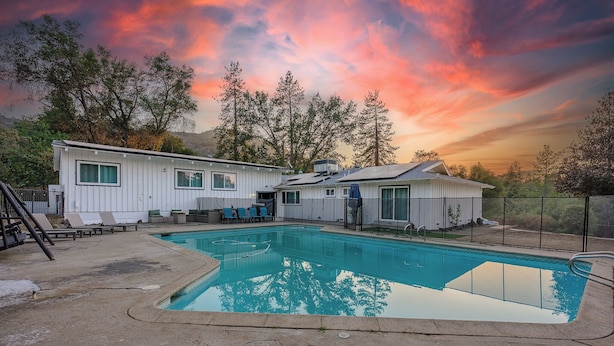 A two-story house with a swimming pool, outdoor seating, and a view of trees.