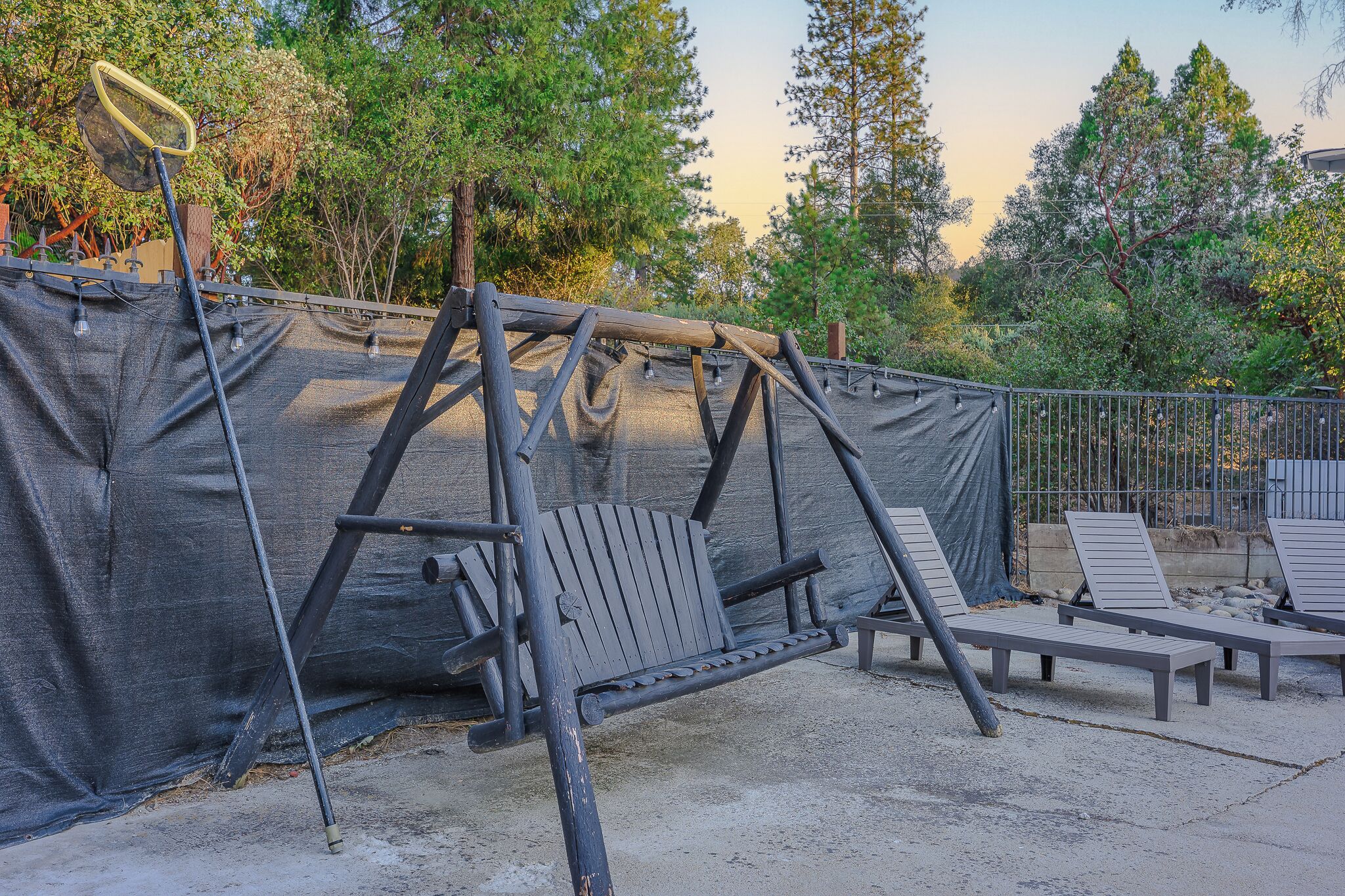 Un conjunto de columpios de madera con red de seguridad, rodeado de árboles y un área cercada con sillas de descanso.