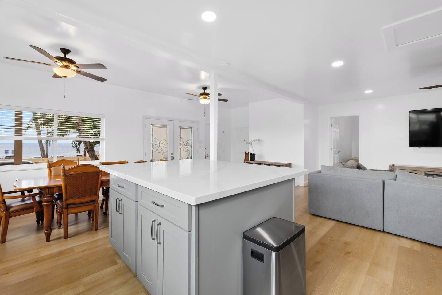 A modern kitchen with a central island, wooden dining table, and chairs, a grey sofa, and a TV mounted on the wall.