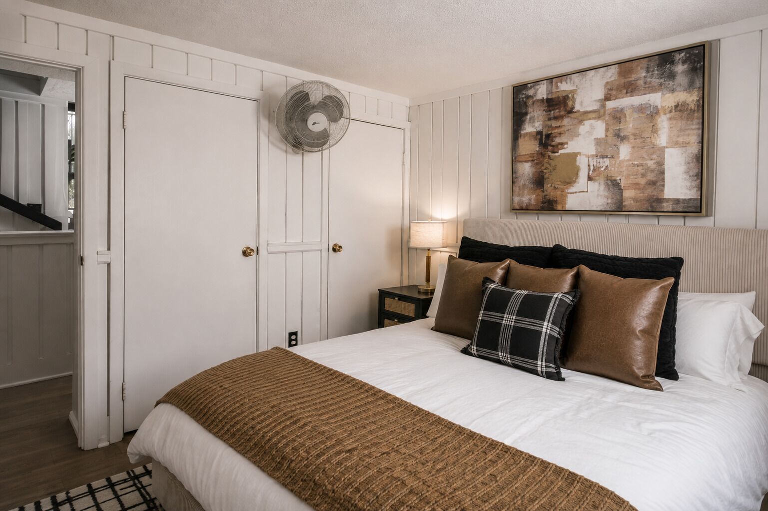 A hotel room with a bed, a bedside lamp, a wall-mounted fan, a door, and a painting.