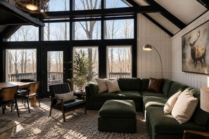 Un salon chaleureux avec un canapé d’angle, une table à manger avec des chaises et une grande fenêtre donnant sur la forêt.