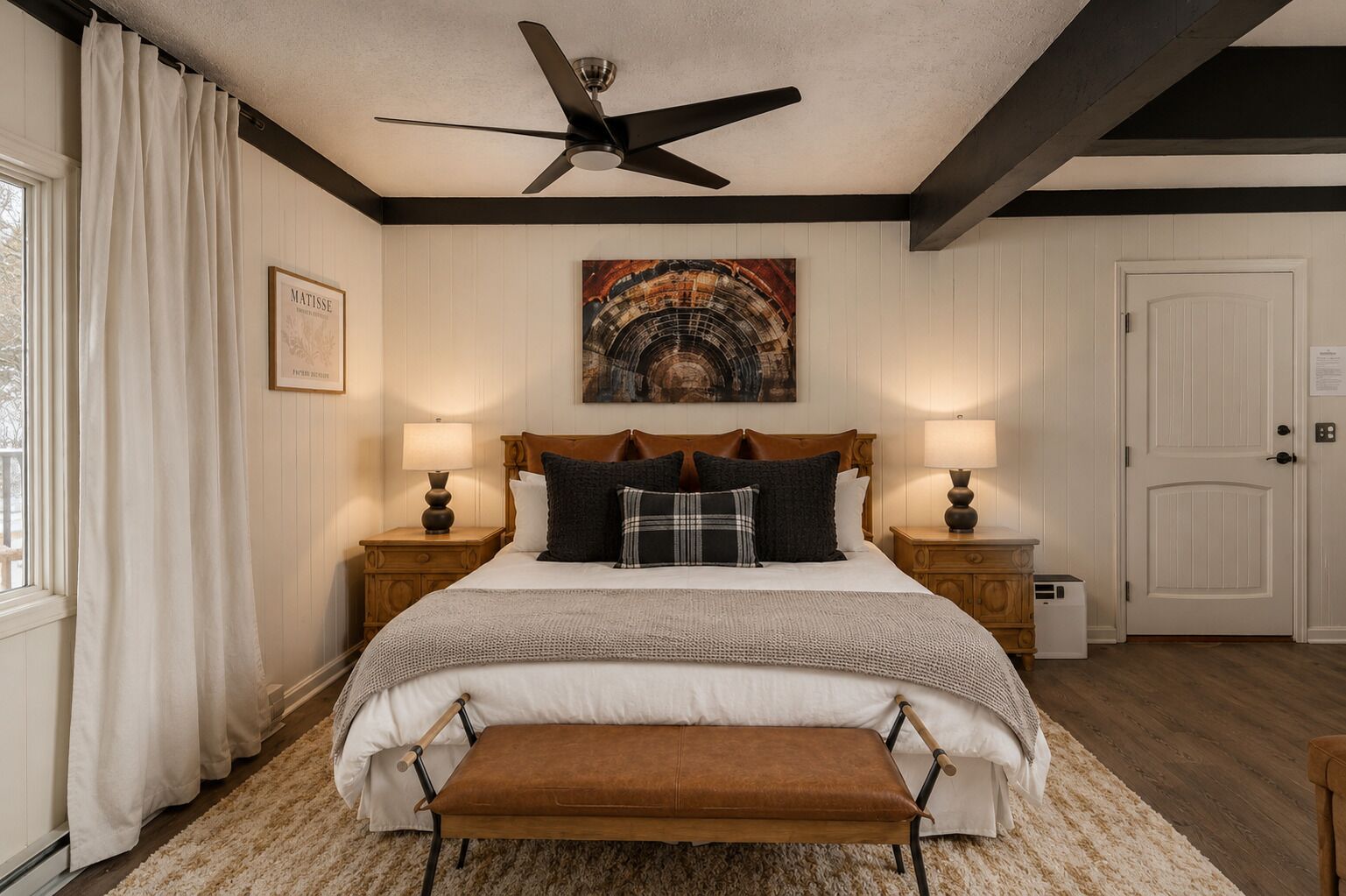 A bedroom with a bed, two bedside tables, a bench, a ceiling fan, a door, and a painting on the wall.