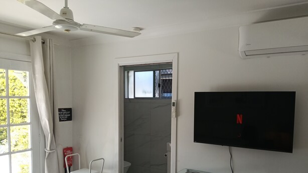 Une chambre d’hôtel comprenant un lit, un ventilateur de plafond, un téléviseur fixé au mur, une table en verre avec une chaise et un espace douche.
