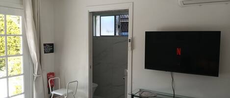 Une chambre d’hôtel comprenant un lit, un ventilateur de plafond, un téléviseur fixé au mur, une table en verre avec une chaise et un espace douche.