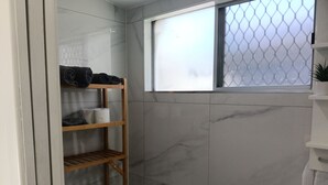 Une salle de bain avec un mur recouvert de carreaux de marbre, une étagère en bois où sont posées des serviettes et une fenêtre munie d’un moustiquaire.