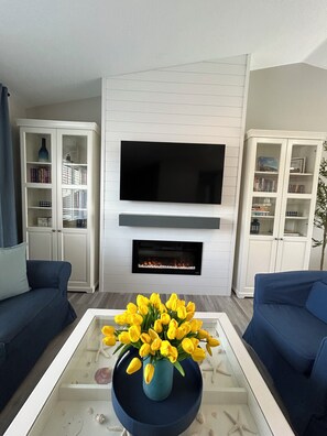 A living room with a flat-screen TV mounted above a fireplace, two blue sofas, and a coffee table with a vase of yellow flowers.