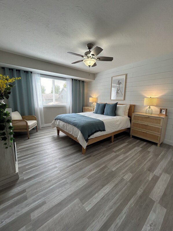 A bedroom with a bed, bedside tables, a ceiling fan, a window with curtains, and a wall-mounted lamp.