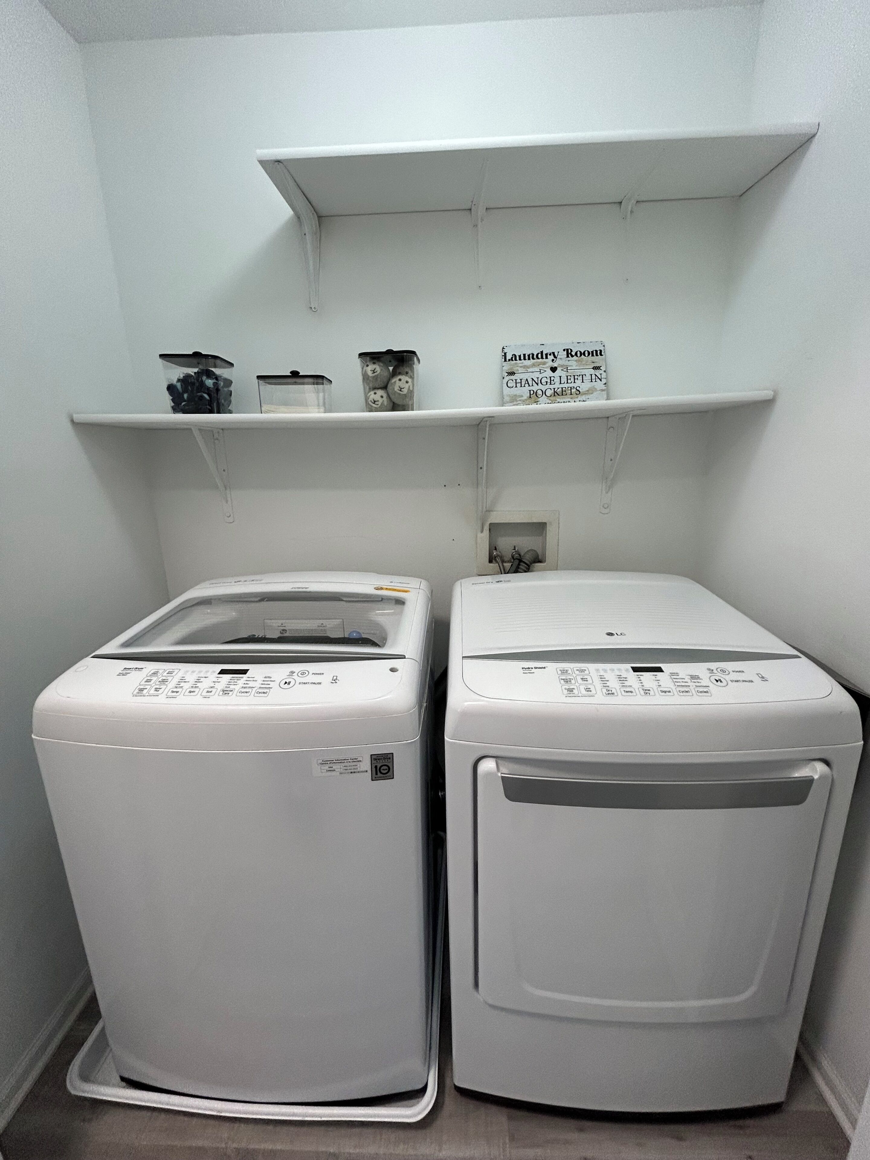 Two front-loading washing machines in a laundry room with a shelf above containing laundry supplies.