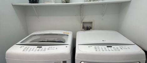 Two front-loading washing machines in a laundry room with a shelf above containing laundry supplies.