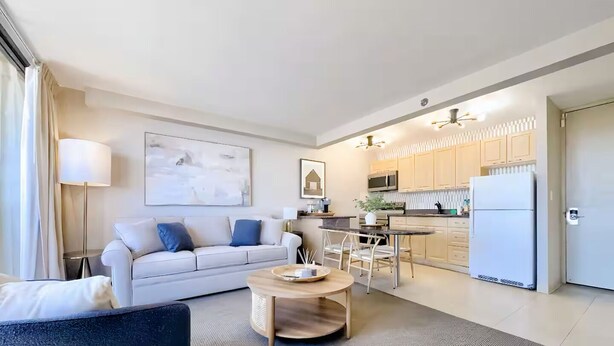Ein modernes Wohnzimmer mit einer Couch, einem Couchtisch und einem Essbereich mit Stühlen. Es gibt eine Küche mit Schränken und einem Kühlschrank.