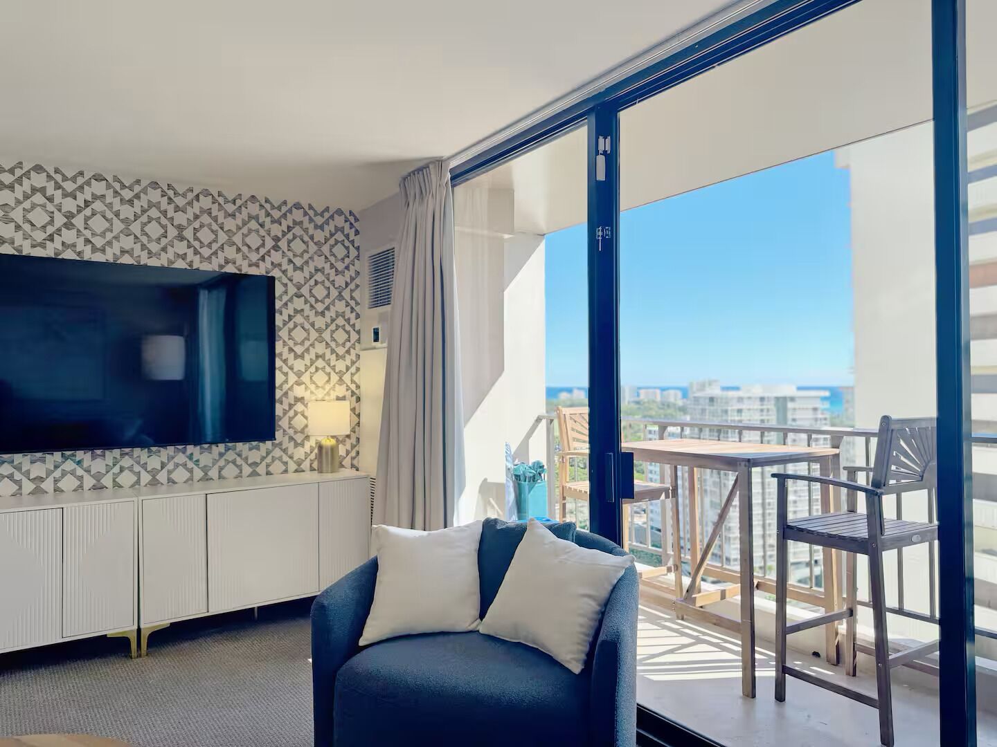 A modern hotel room with a large window, a flat-screen TV, a sofa, and a balcony with a table and chairs.