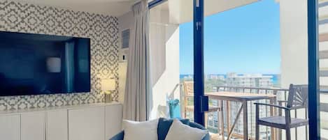 A modern hotel room with a large window, a flat-screen TV, a sofa, and a balcony with a table and chairs.