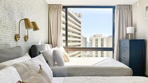 Ein Hotelzimmer mit zwei Betten, einem großen Fenster mit Blick auf Gebäude, einem Kleiderschrank und einer Lampe.