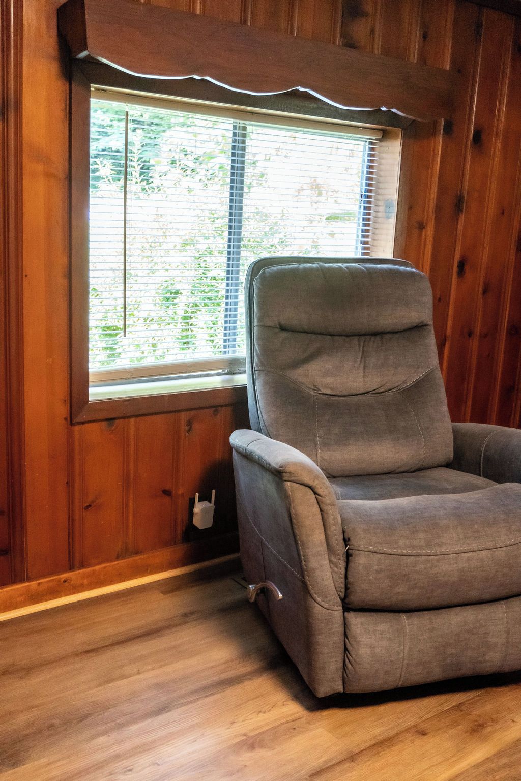 Una habitación con un revestimiento de madera, una ventana con persianas y un sillón tapizado en gris.