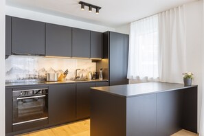 A modern kitchen with dark cabinetry, a built-in oven, and a large island. - Ofs_c_fw_2sz_m.t._mh_fis_13 (Fiss)