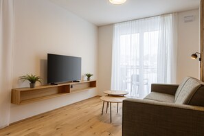Ein modernes Wohnzimmer mit einem Flachbildfernseher auf einem Holzbodenregal, einem grauen Sofa, einem kleinen runden Tisch und zwei Blumentöpfen.